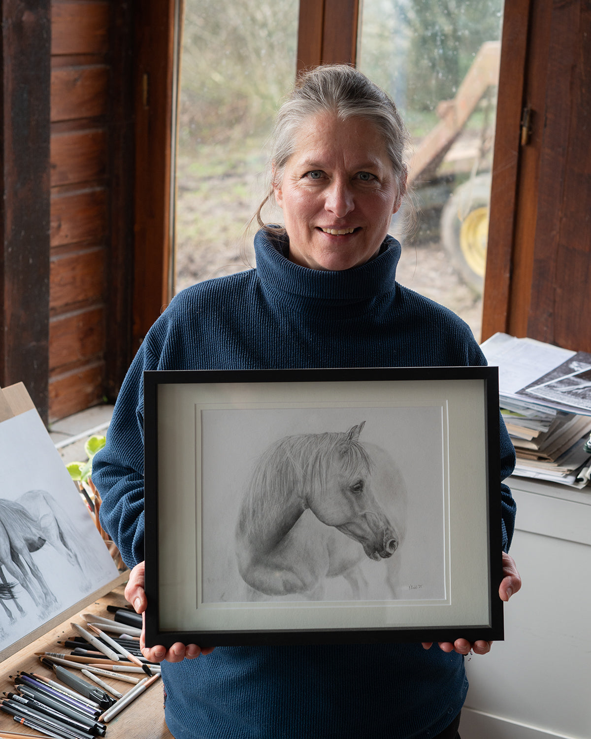 Person holding a framed drawing of a horse in a room with wooden walls and a window.